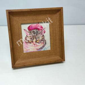 以下が含まれることがあります： ピンクのベレー帽をかぶった不機嫌そうな猫の水彩画の額入り。猫は黄色い目をし、厳しい表情をしています。フレームはミディアムブラウン仕上げの木製です。「moxx kit」の文字が一部表示されています。