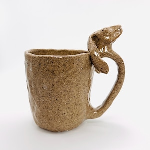 May include: A handmade ceramic mug with a unique dog-shaped handle. The mug is a speckled brown color with a textured surface. The dog handle adds a whimsical touch to this functional piece.