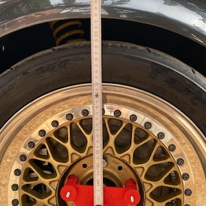 Puede incluir: Primer plano de una rueda de coche con una llanta dorada y un neumático negro. Se coloca una herramienta de medición roja en la rueda y una regla se coloca verticalmente. La carrocería gris del coche es visible encima de la rueda.