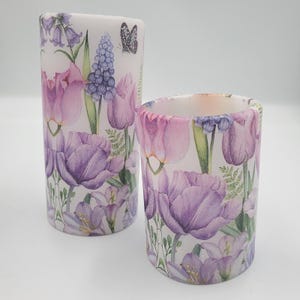Könnte beinhalten: Zwei zylindrische Kerzen mit floralen Mustern. Die größere Kerze zeigt lila Tulpen, Glockenblumen und einen Schmetterling. Die kleinere Kerze hat ein ähnliches Design. Beide Kerzen haben einen weißen Hintergrund und sind mit grünem Laub verziert.