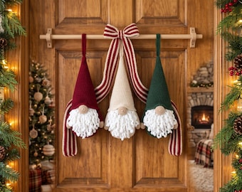 May include: Three crocheted gnomes hanging from a wooden rod, featuring pointed hats in dark red, cream, and dark green. Each gnome has a round beige nose and a thick, white, curly yarn beard. A striped red and white ribbon bow connects them.