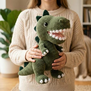 May include: A green crocheted dinosaur toy with white teeth and a pink mouth. The dinosaur has dark green spikes and white accents on its feet and hands. The toy is held in front of a person wearing a cream-coloured jumper.