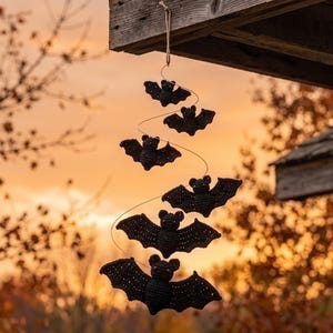 May include: A hanging mobile featuring five black crocheted bats of varying sizes arranged in a spiral pattern. The bats are suspended from a thin wire attached to a piece of wood by a short length of twine, creating a vertical display.