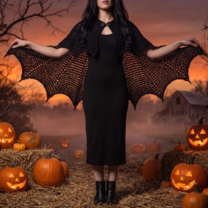 May include: A black crochet shrug featuring a bat wing design with scalloped edges and a single button closure at the neckline, worn over a simple black sleeveless dress, creating a distinct silhouette with pointed wing-like extensions.