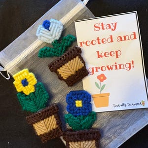 May include: Handmade, colorful, crocheted flower magnets in brown pots with green leaves. The flowers are blue, yellow, and light blue. A card reads "Stay rooted and keep growing!" with a flower illustration. The magnets are in a sheer bag.