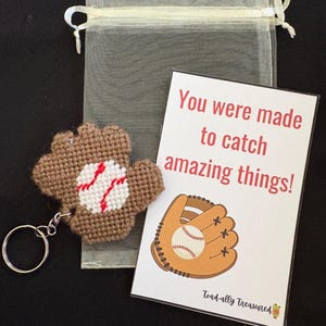 May include: A baseball-themed gift set featuring a brown and white baseball glove keychain, a sheer drawstring bag, and a card with the text "You were made to catch amazing things!" and a baseball glove illustration.