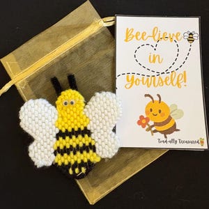 Puede incluir: Un broche artesanal con forma de abeja en amarillo, negro y blanco, junto a una tarjeta con el mensaje "¡Cree en ti mismo!" y una ilustración de abeja. El broche y la tarjeta se presentan con una bolsa de regalo de malla dorada.