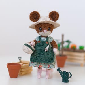 Könnte beinhalten: Eine braune gehäkelte Mausfigur mit beigem Sonnenhut, einem hellgrünen kurzärmeligen Kleid mit Blumenmuster und einer dunkelgrünen gehäkelten Schürze. Die Maus hält einen kleinen beigen Korb und hat eine silberne Kelle in der Schürzentasche.
