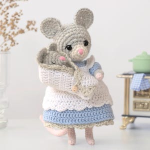 Puede incluir: Una muñeca de ratón tejida a ganchillo de color gris con ojos negros y nariz rosa, que lleva un vestido azul claro con un delantal blanco. El ratón sostiene un bebé ratón gris envuelto en una manta blanca con un botón y borde beige.