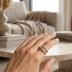 Puede incluir: Un anillo de oro con un diseño único que presenta múltiples bandas entrelazadas, algunas adornadas con pequeñas piedras brillantes. El anillo se muestra en una mano, mostrando sus intrincados detalles y su estilo elegante. El anillo es una joya.