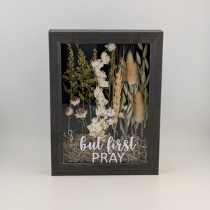 May include: A rectangular framed art piece featuring dried flowers and the text "but first PRAY". The frame is dark gray, and the flowers are white, beige, and green, set against a black background.