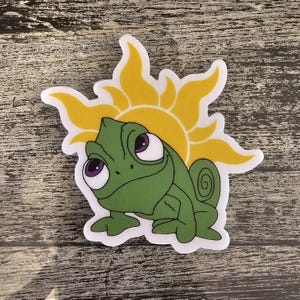 Op de afbeelding: Een groene kameleon met paarse ogen en een gele zon erachter. De kameleon kijkt naar rechts en heeft een gekrulde staart.