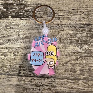 May include: A clear acrylic keychain featuring a cartoon character with yellow skin, a white collar, and a wide grin. The keychain has Japanese text and a pink and white design. A gold-colored key ring is attached.