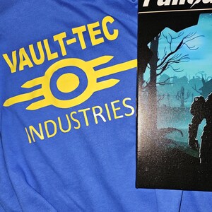 May include: A blue long-sleeve shirt with the words "VAULT-TEC INDUSTRIES" in yellow, along with the Vault-Tec logo. Next to the shirt is a canvas print with the word "Fallout" and a post-apocalyptic scene.