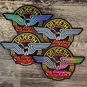 Könnte beinhalten: Vier schwarze Aufkleber mit einem kreisförmigen Design mit dem Text "DR. TEETH and The Electric Mayhem". Das Design umfasst einen Zahn mit Zylinder und stilisierte Flügel in Blau-, Gelb- und Rottönen. Die Aufkleber haben ein holografisches Finish.