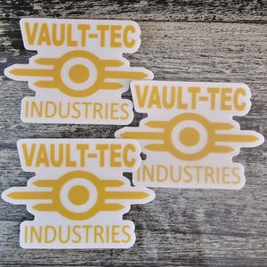 May include: Three white stickers featuring the Vault-Tec Industries logo in yellow. The logo has a central circle with horizontal lines extending outwards, and the words "VAULT-TEC" and "INDUSTRIES" are printed above and below.