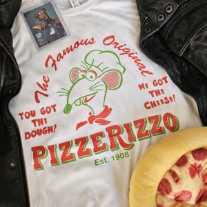 May include: White t-shirt with a cartoon rat chef graphic and the text "PIZZERIZZO Est. 1908". The shirt also features the phrases "The Famous Original", "You Got The Dough?", and "We Got The Cheese!". A black leather jacket and a pizza-shaped pillow are also in the image.