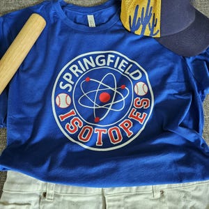 May include: A royal blue t-shirt with a white and red graphic of a baseball and an atom design, with the words "Springfield Isotopes". A baseball bat and a navy blue baseball cap with yellow accents are also visible.