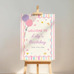 May include: A rectangular birthday welcome sign on a wooden easel featuring a cream-colored center with a scalloped pink border, pastel striped edges, purple and pink balloons, and the text: Welcome to Lily's Birthday, June 14th, 2026.