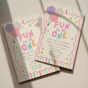 Op de afbeelding: Twee rechthoekige verjaardagsuitnodigingskaarten met pastelkleurige verticale strepen en een geschulpte rand. De tekst luidt FUN to be ONE, Lily's First Birthday, Sunday, June 14 at 2PM, Our Place, 126 Lovely Lane, RSVP by 6/10, 123.456.7890.