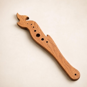 May include: A handcrafted wooden bottle opener with a unique, flame-like design. The opener is made of a warm, reddish-brown wood and features several circular cutouts along its curved body. The design includes a small hole at the end for hanging.