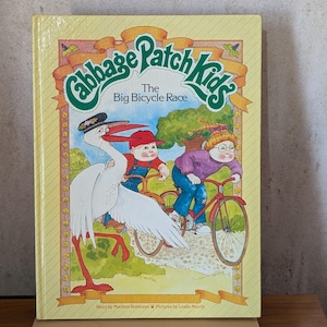 May include: A hardback children's book titled Cabbage Patch Kids The Big Bicycle Race. The yellow cover features an illustration of a large white stork wearing a hat, and two children riding bicycles on a path. The text includes the author and illustrator names.