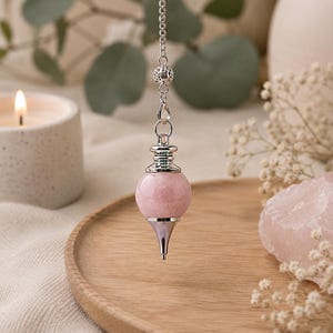 May include: A dowsing pendulum featuring a smooth, spherical light pink stone bead attached to a silver-toned metal cap and a pointed silver-toned metal tip. The pendulum hangs from a delicate silver-toned metal chain with a small decorative bead.