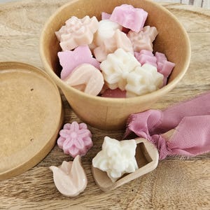 Può includere: Un contenitore rotondo in carta color cuoio riempito con piccole forme di cera modellate nei toni del bianco, crema e rosa chiaro. Le forme includono fiori e boccioli di tulipano. Un piccolo cucchiaio di legno con un nastro rosa polvere contiene una cera.