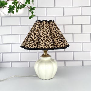 Puede incluir: Una lámpara de mesa decorativa con una base de color crema y una pantalla con estampado de leopardo. La pantalla tiene un ribete negro y un borde festoneado. La lámpara tiene un cuello color latón y un cable blanco.