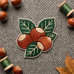 May include: An embroidered patch featuring three brown hazelnuts with light tan tops, surrounded by dark green leaves with white vein detailing. The patch has a cream-colored border and is displayed on a textured gray fabric background.