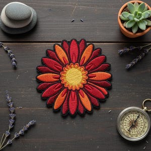 Könnte beinhalten: Ein kreisförmiger gestickter Aufnäher, der eine stilisierte Blume mit einem leuchtend gelben Zentrum zeigt, umgeben von abwechselnden Blütenblättern in tiefen Rot- und leuchtenden Orangetönen, eingefasst von einem dünnen schwarzen Rand.