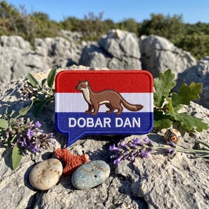 May include: A rectangular embroidered patch shaped like a speech bubble with a red, white, and blue horizontal striped background. A brown weasel is centred on the patch above the white text DOBAR DAN printed in a bold, sans-serif font.