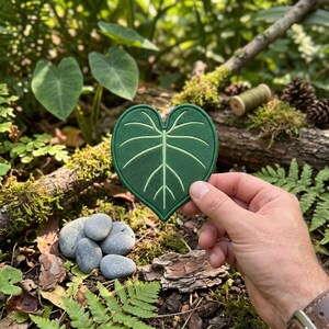 May include: A heart-shaped patch featuring a dark green base with embroidered light green veins, mimicking the appearance of a leaf. The patch has a stitched border and is held up against a natural outdoor background of foliage and wood.