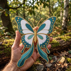 May include: A butterfly-shaped embroidered patch featuring teal and white thread with gold-colored metallic thread outlining the wings and body. The design includes intricate stitching patterns and elongated lower wing sections with a symmetrical layout.