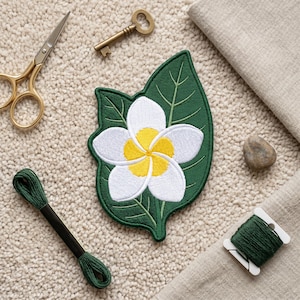 May include: An embroidered patch featuring a white plumeria flower with a yellow center, set against a dark green leaf background. The patch is surrounded by gold scissors, a brass key, a small stone, and dark green embroidery thread.