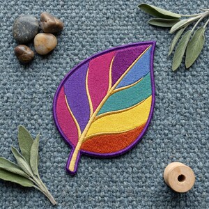 Pode incluir: Um emblema bordado em forma de folha com segmentos vibrantes em roxo, magenta, azul-petróleo, azul, amarelo e laranja, contornados por uma borda roxa escura. O design inclui um caule central amarelo e nervuras que separam cada seção colorida.