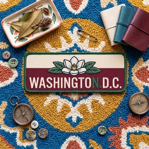Puede incluir: Un parche rectangular bordado que presenta una flor de magnolia blanca con hojas verdes sobre el texto WASHINGTON D.C. en letras blancas y verdes, sobre un fondo horizontal crema y granate con un borde de color verde.
