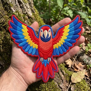 Peut inclure: Un écusson brodé en forme de perroquet ara aux ailes largement déployées. L'oiseau présente un corps rouge vif, des plumes d'ailes jaunes et bleues, un bec crème et une tache bleu foncé autour des yeux, le tout bordé d'un contour rouge vif.