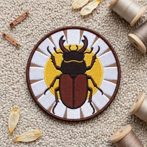May include: A circular embroidered patch featuring a brown stag beetle centred on a yellow circle, surrounded by a white and brown sunburst pattern. The patch has a dark brown stitched border and is displayed with sewing supplies.