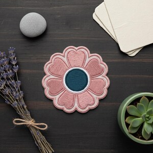 May include: A circular embroidered patch featuring a stylized flower design with dusty pink petals, a dark teal center, and a white border. The patch is displayed alongside a smooth gray stone, a stack of cream paper, dried lavender, and a green succulent.