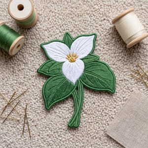 May include: An embroidered patch featuring a white trillium flower with three petals, a yellow center, and dark green leaves. The patch is surrounded by two wooden spools of green and white thread, several sewing needles, and a piece of fabric.