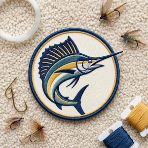 May include: A circular embroidered patch featuring a blue and yellow sailfish on a cream background with a dark blue border. The patch is surrounded by fishing gear including four feathered lures, a spool of clear line, and two spools of thread.