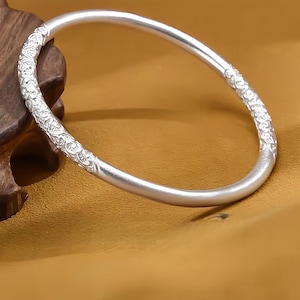 May include: A silver bangle bracelet with decorative floral patterns. The bracelet has a smooth, polished finish with detailed patterns on the ends. The bracelet is curved and rests on a wooden surface.