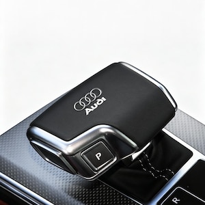 May include: A black leather-wrapped Audi gear shifter with a metallic silver frame and a prominent P button on the side. The top surface features the silver Audi logo and text. The base includes a black leather boot with white stitching and gear indicators.