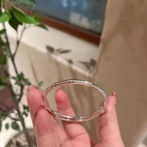 Op de afbeelding: Een zilverkleurige armband met een bamboe-ontwerp. De armband wordt in een hand gehouden, waarbij de gepolijste afwerking en de gesegmenteerde structuur worden benadrukt. Het sieraad is een bangle-stijl, met een glad, reflecterend oppervlak.