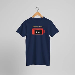 Könnte beinhalten: Marineblaues T-Shirt mit einer rot-schwarzen Grafik einer Batterie mit 1 % Ladung. Der Text "ENERGY LEVEL" befindet sich über der Grafik. Das T-Shirt hängt an einem Holzbügel.