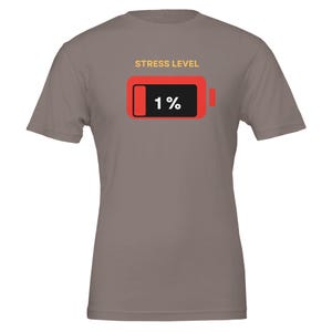 Könnte beinhalten: Taupefarbenes T-Shirt mit der Aufschrift "STRESS LEVEL" über einer roten Batterie-Grafik mit "1%" in Weiß. Das Shirt hat kurze Ärmel und einen Rundhalsausschnitt.