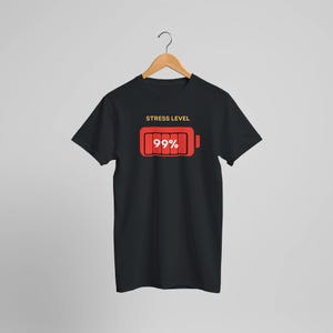 Könnte beinhalten: Schwarzes T-Shirt mit roter Batterie-Grafik, die "99%" und die Worte "STRESS LEVEL" zeigt. Die Batterie ist fast voll, was auf hohen Stress hindeutet. Das T-Shirt hängt an einem Holzkleiderbügel.