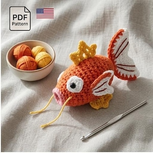 Puede incluir: Un pez tejido a ganchillo en naranja y blanco con detalles amarillos y boca rosa. Un cuenco pequeño con ovillos de hilo naranja y amarillo está junto al pez. También se ven un ganchillo y un icono de patrón PDF.