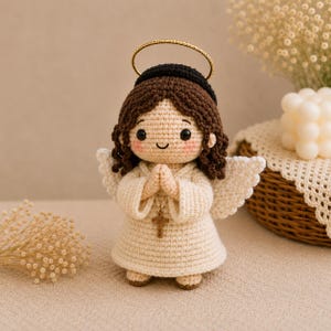 May include: A crocheted angel doll with dark brown hair, a gold halo, and a cream-colored robe. The doll has black eyes, pink cheeks, and hands pressed together in prayer. A small wooden cross pendant hangs from a string around the doll's neck.